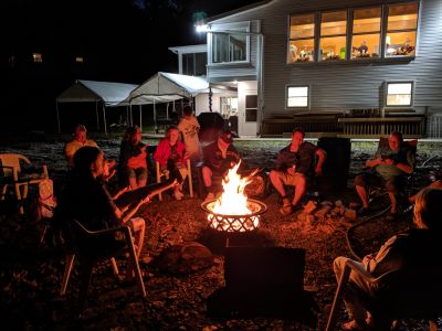 60th Anniversary Party Jack & Jan August 10, 2019
Campfire 6 
pic by Joe.  
Note the people in the Great Room, and the outside setup we had to accommodate everyone either indoors or outdoors.
L to R: Riley, toasting his "dogs: Jim; Joan; Grandma; Cole; Grandpa; Arik; John; others not in view (see other camp fire shots); Ann
